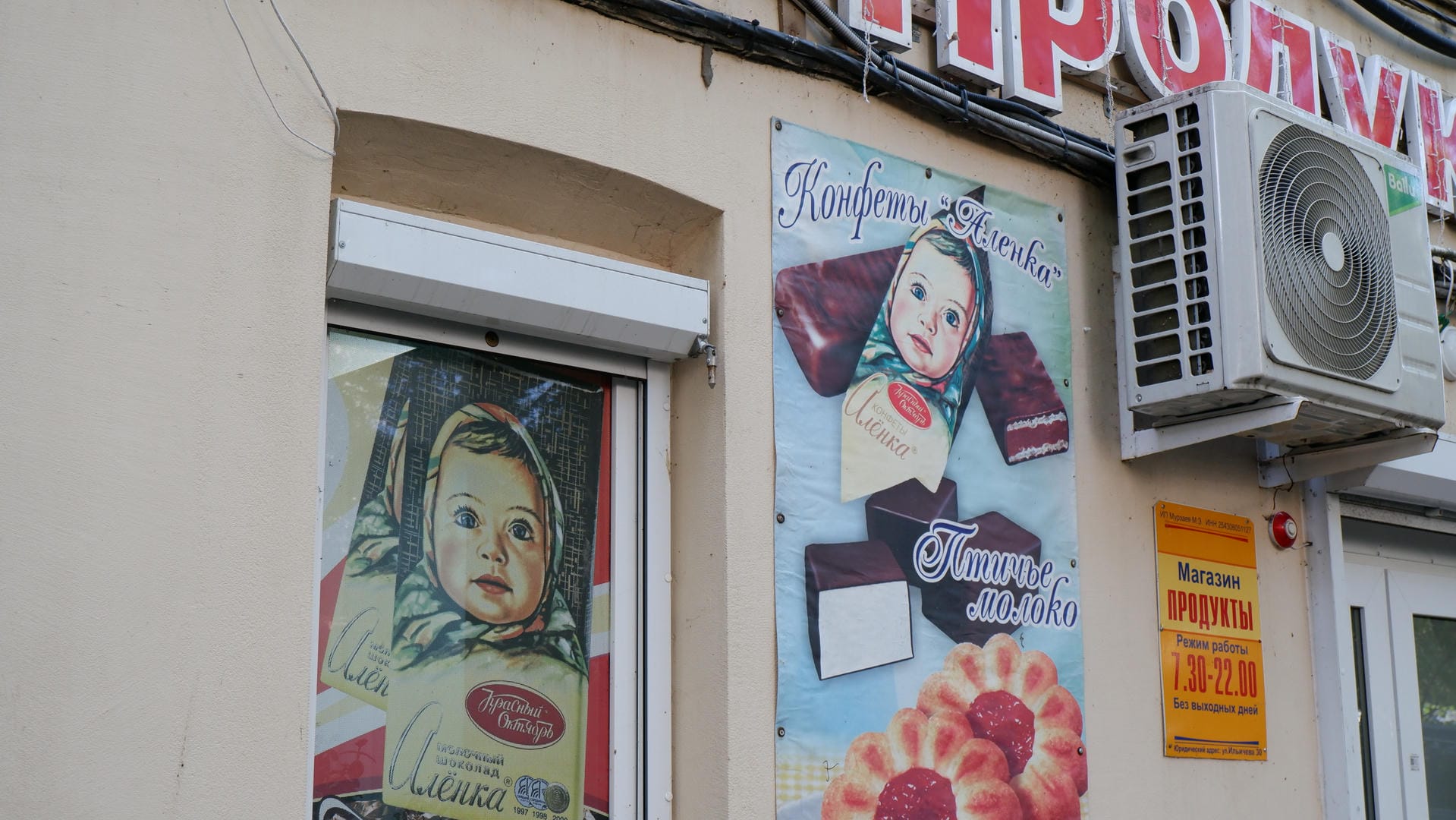 An advertisement for baby food is posted on the exterior wall of a store, with an air conditioning unit hanging nearby.