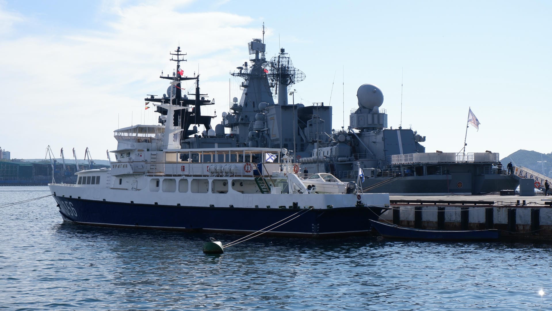 A white and blue boat docked at a pier with a large warship in the background.