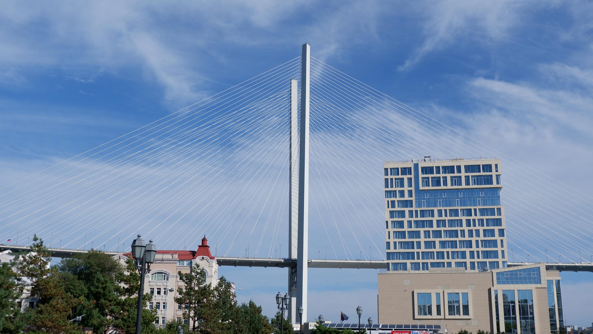 The tower of a suspension bridge reaching into the clouds, next to modern buildings and greenery.