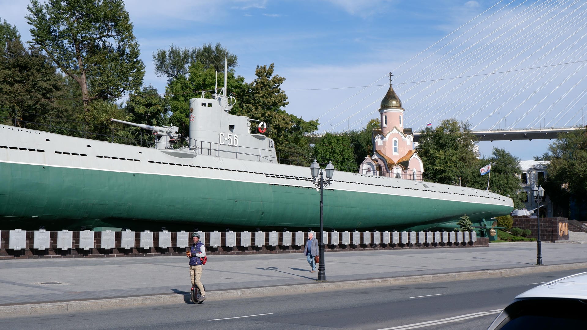 A green submarine displayed in a park, next to a church and a person riding a unicycle.