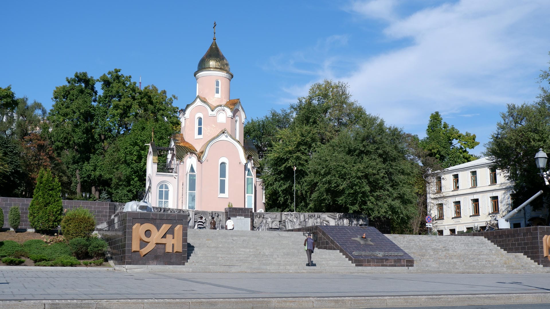 A pink church with a memorial plaque from 1941 in front, surrounded by green trees and sculptures.