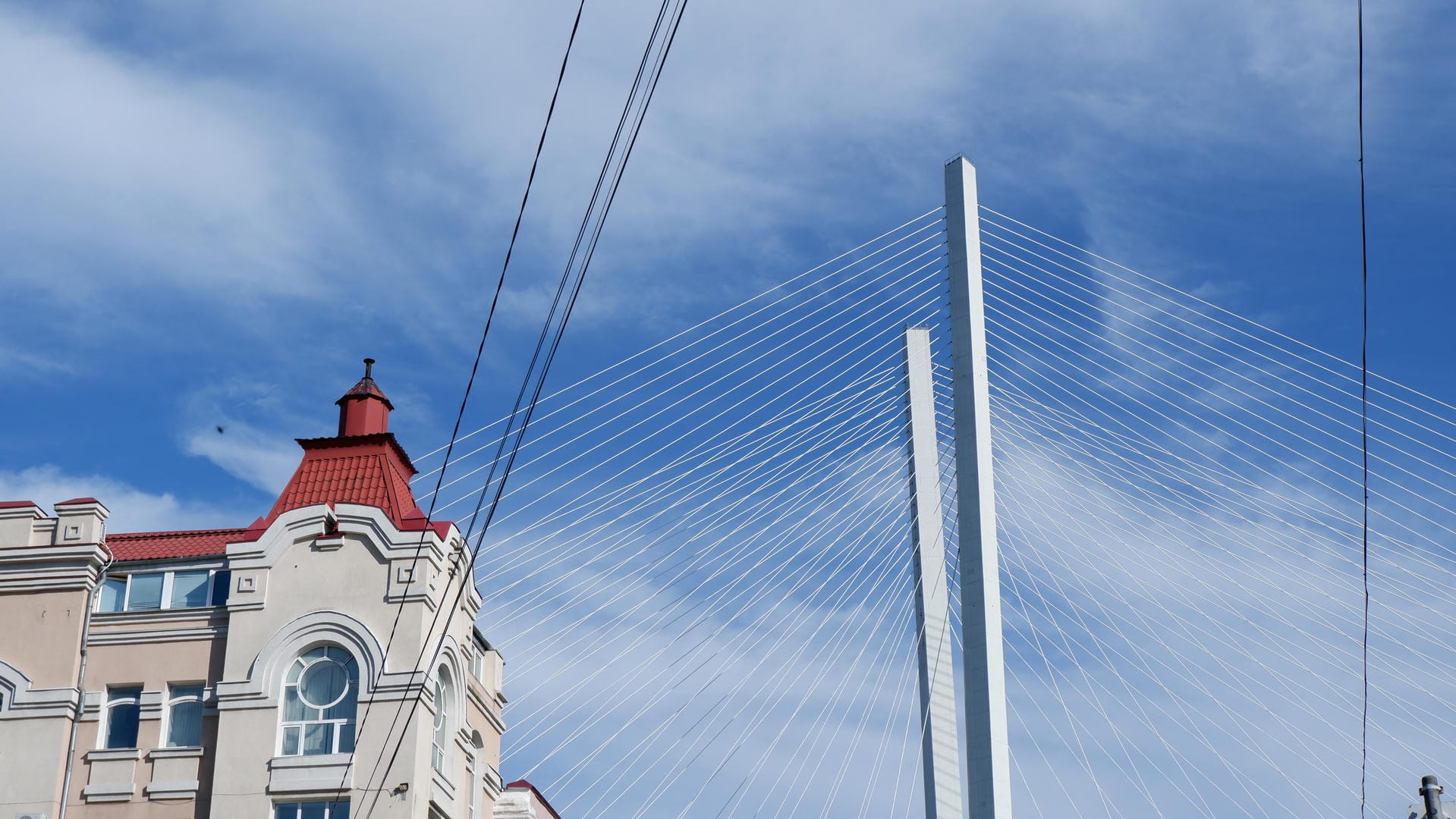 The tower of a suspension bridge reaching into the clouds, next to a red-roofed building.