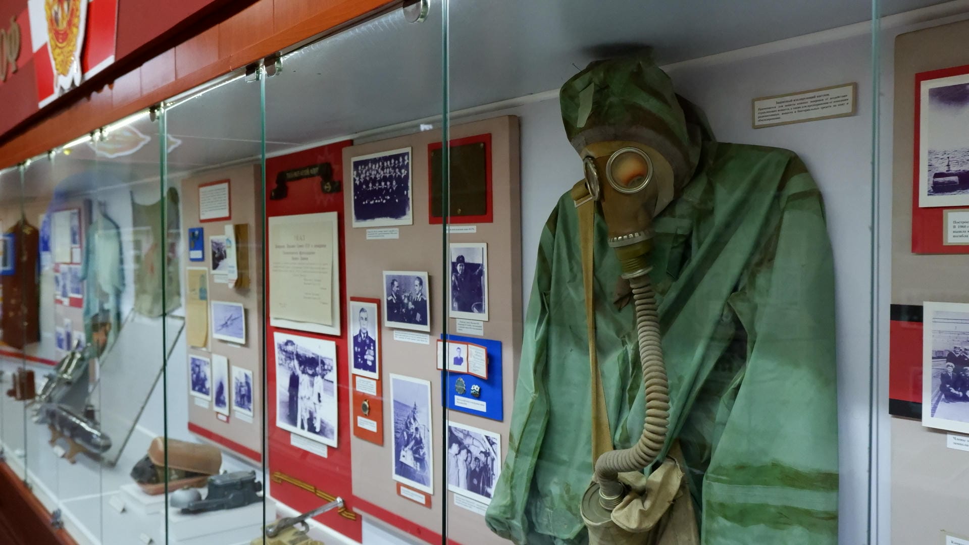 A protective suit and gas mask hanging on the wall, next to photographs and documents.