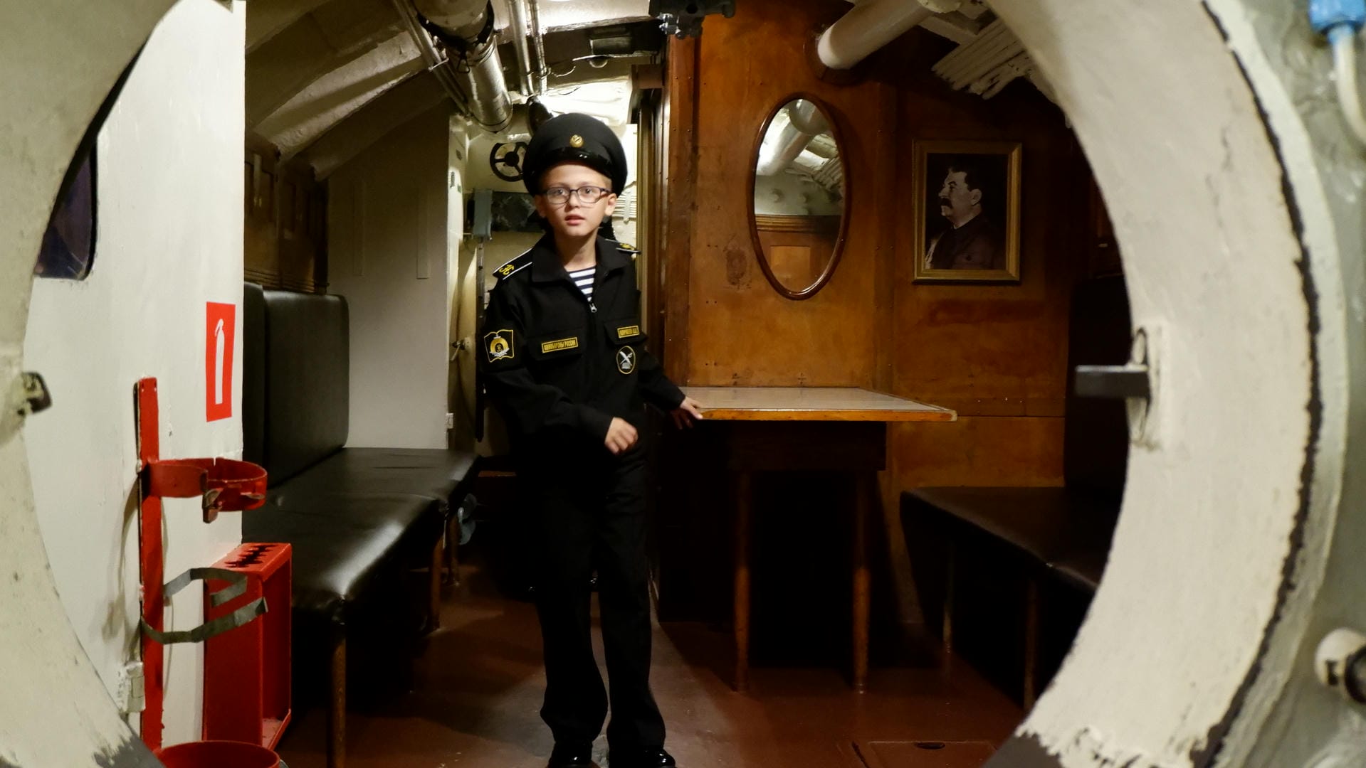 A young boy dressed in naval uniform stands inside the cabin, with a portrait hanging on the wall in the background.