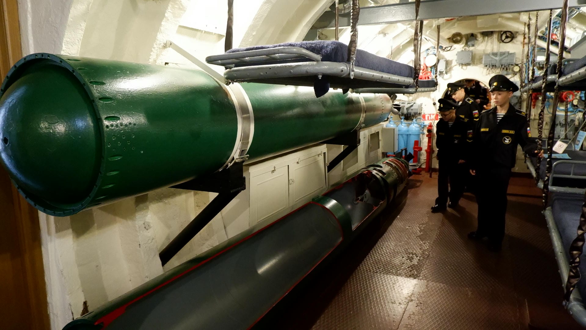 Green torpedoes and other equipment hang on the walls inside the submarine, with several people observing around.