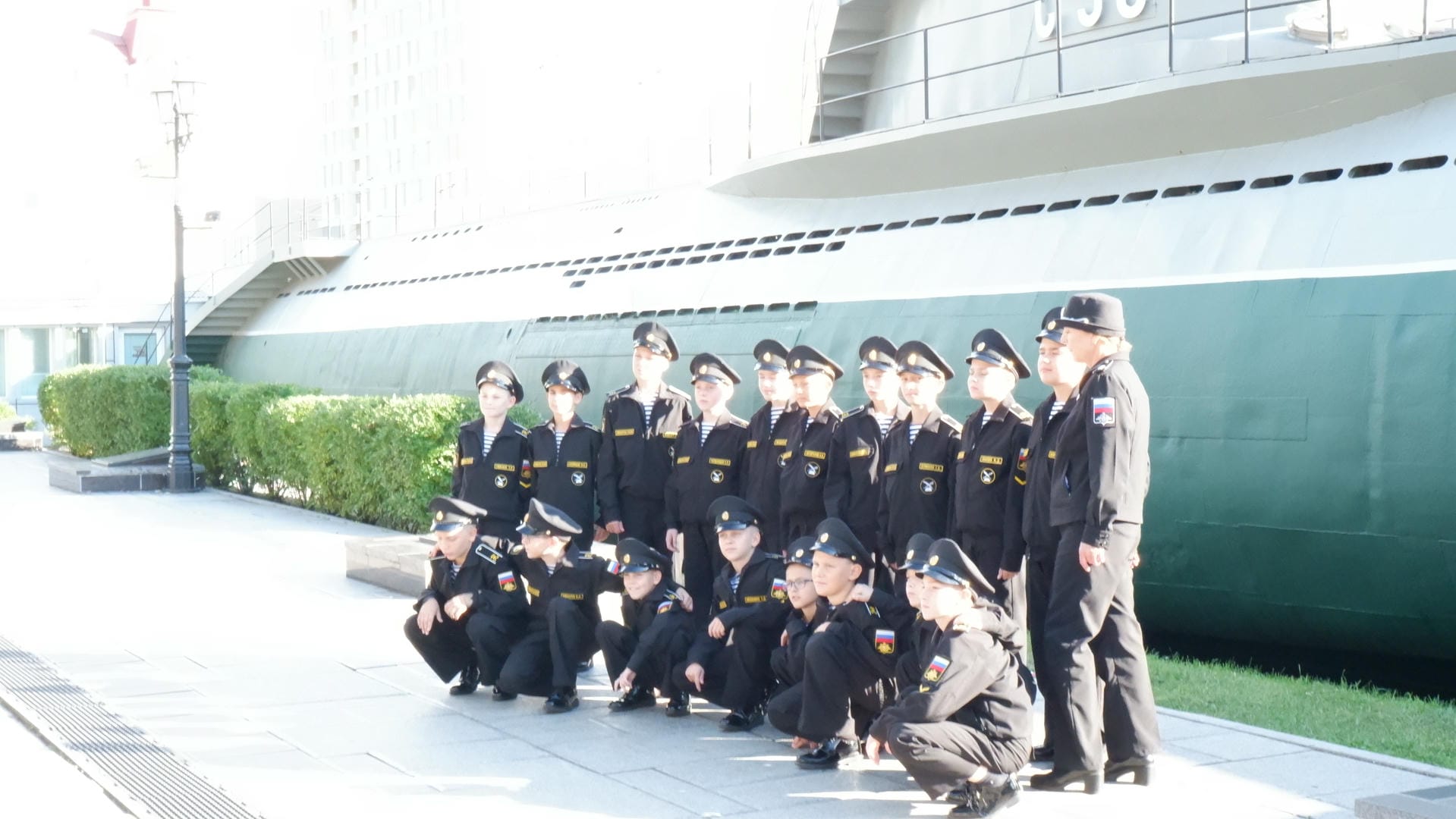 A group of children dressed in naval uniforms stands in front of a submarine, with a clear sky in the background.