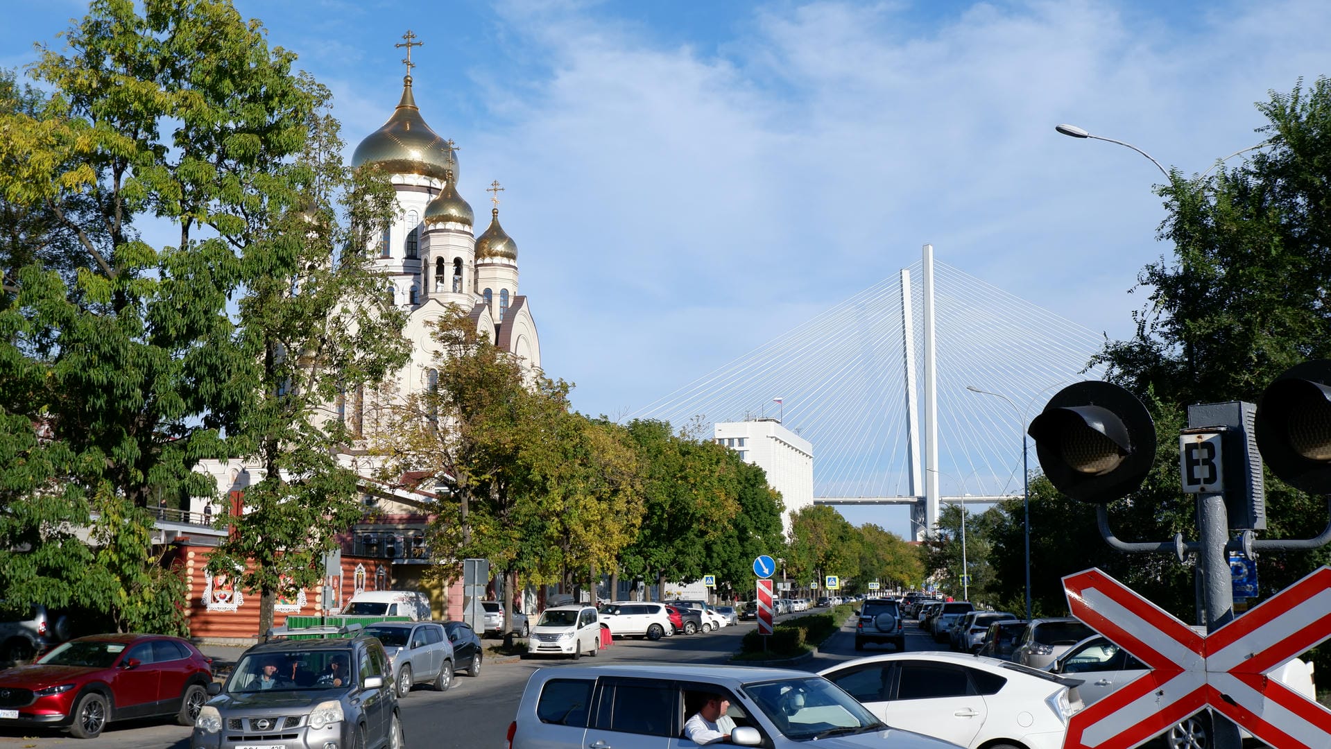 A large submarine is docked by the shore, surrounded by trees and buildings, with a suspension bridge in the distance.