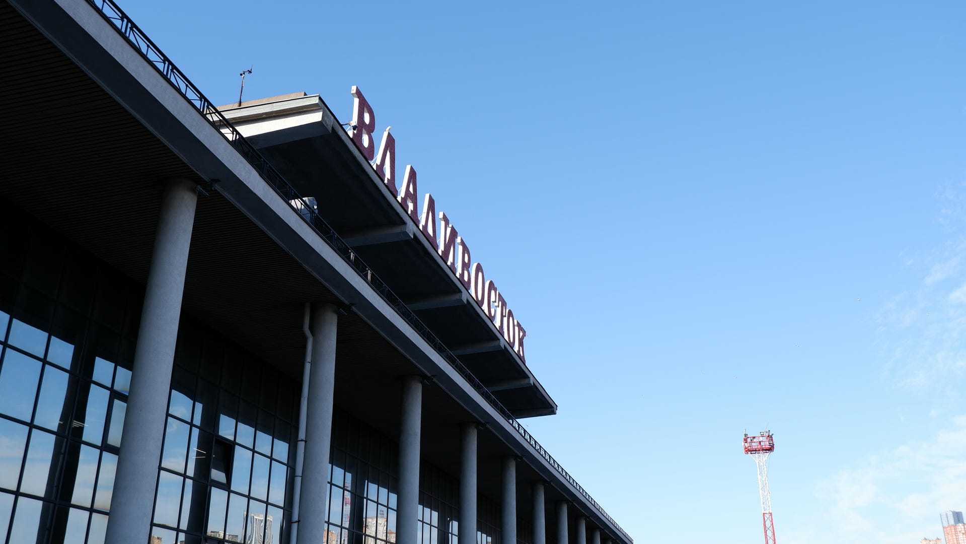 The top of a modern building features large red letters spelling “ВЛАДИВОСТОК”, against a backdrop of blue sky.