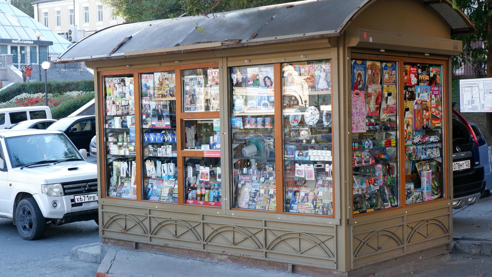A roadside newsstand filled with various magazines and newspapers, with cars parked outside.