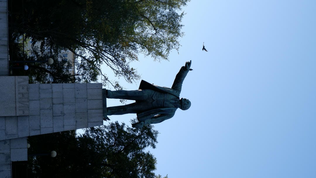 A Lenin statue pointing into the distance stands in a square, surrounded by trees.