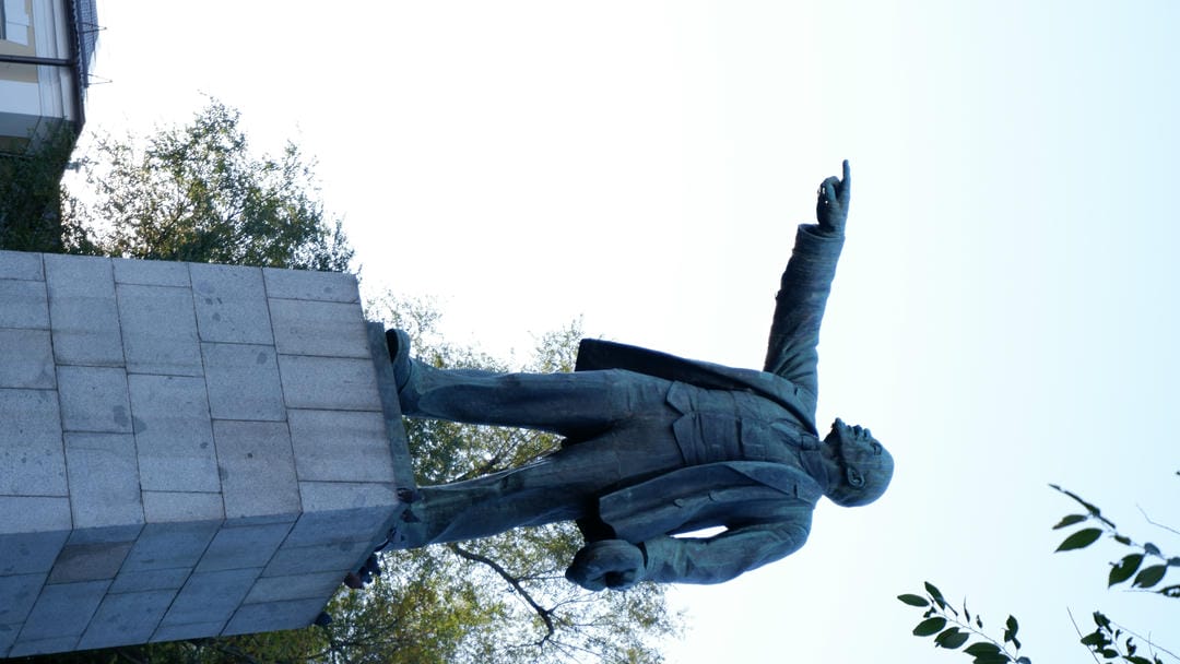 A Lenin statue pointing into the distance stands in a square, surrounded by trees.