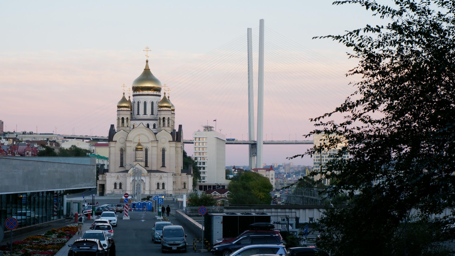 A church with a golden dome, with multiple cars parked on the road ahead, and a suspension bridge in the distance.