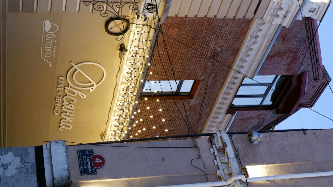 The exterior wall of a building is hung with many small light bulbs, with Russian text signs, and a blue road sign nearby.