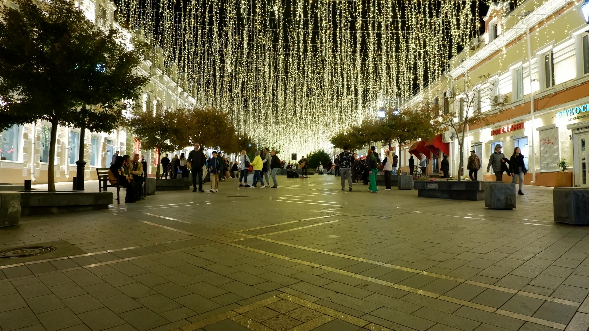 A street decorated with yellow lights, flanked by trees and buildings, with people strolling or sitting.