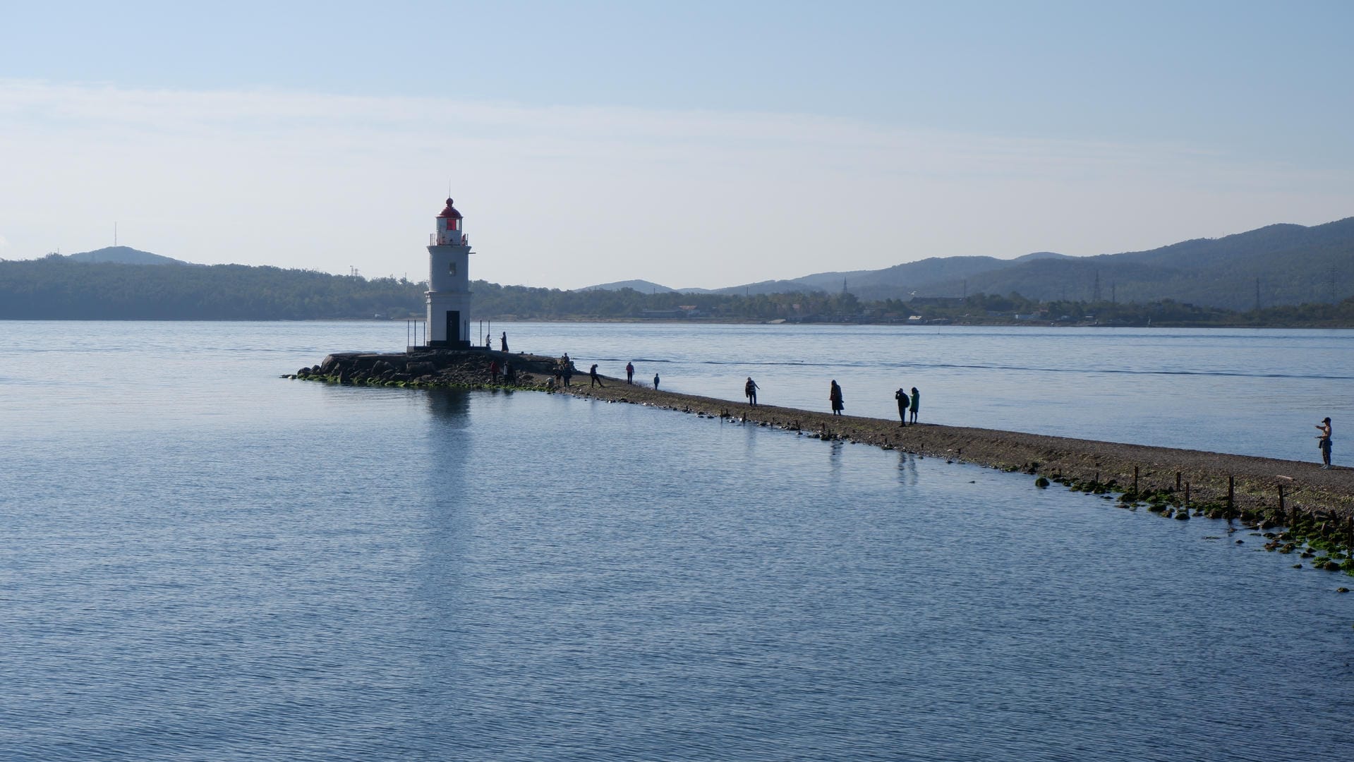 A stone pier extending into the water has a lighthouse, with some people walking around.