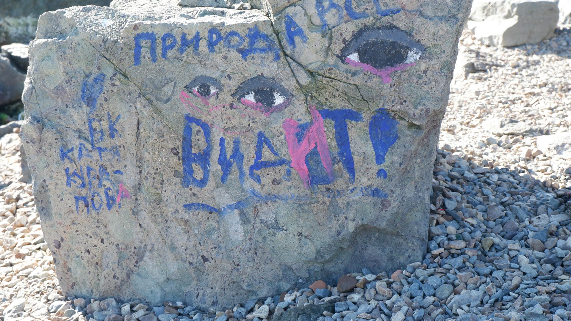 A rock is painted with a pair of eyes and Russian text, surrounded by scattered pebbles.