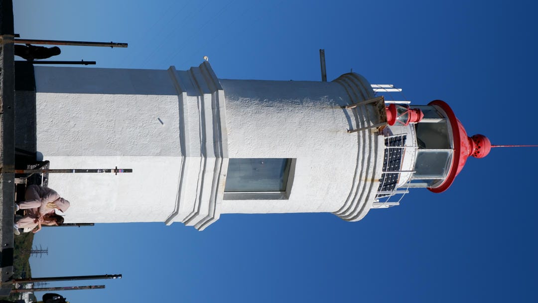 A white lighthouse with a red top, with two people sitting on a bench nearby.