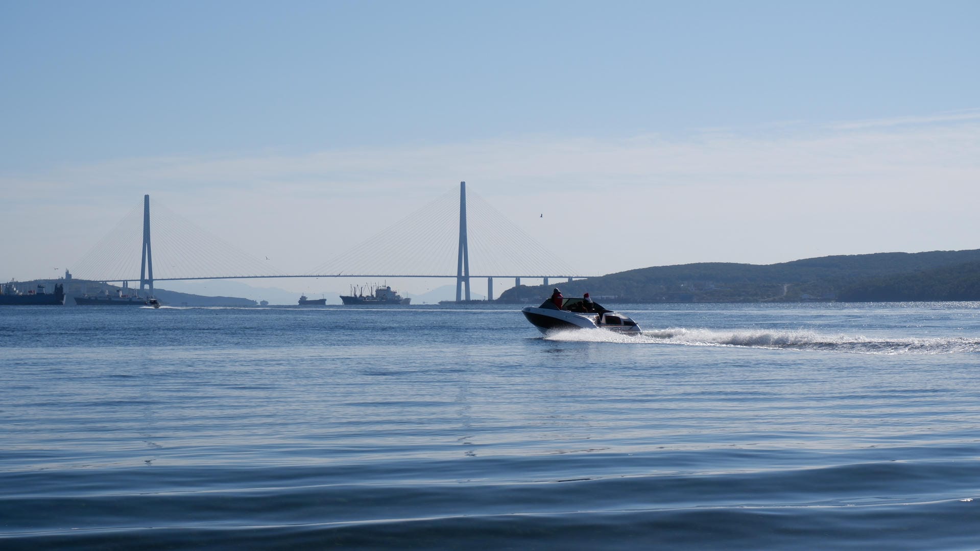 A speedboat travels across the water, with a suspension bridge in the distance and vehicles passing on it.