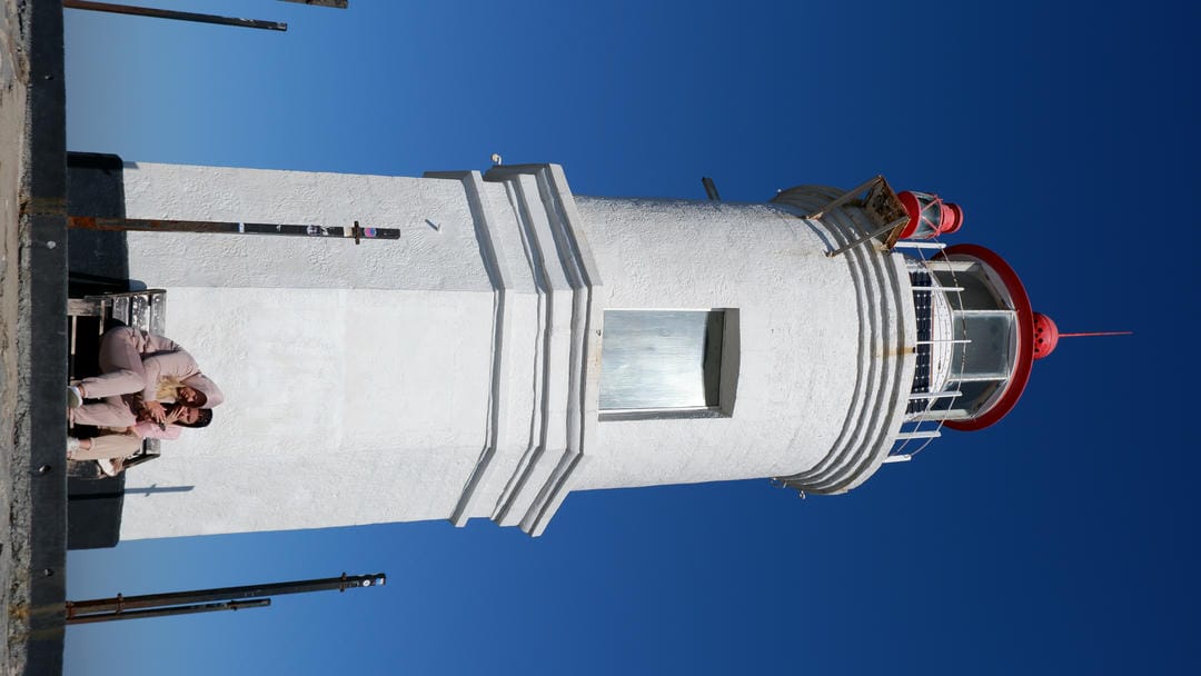 A white lighthouse with a red top, with two people sitting on a bench at the base of the lighthouse.