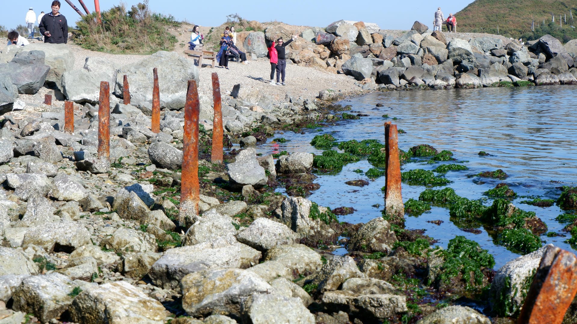 People stand by the rocky coastline, with rusty metal posts in the foreground and seated figures in the background.