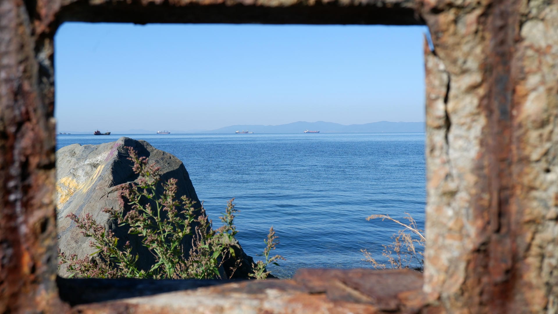 A body of water seen through a rusty square frame, with boats and mountains in the distance.