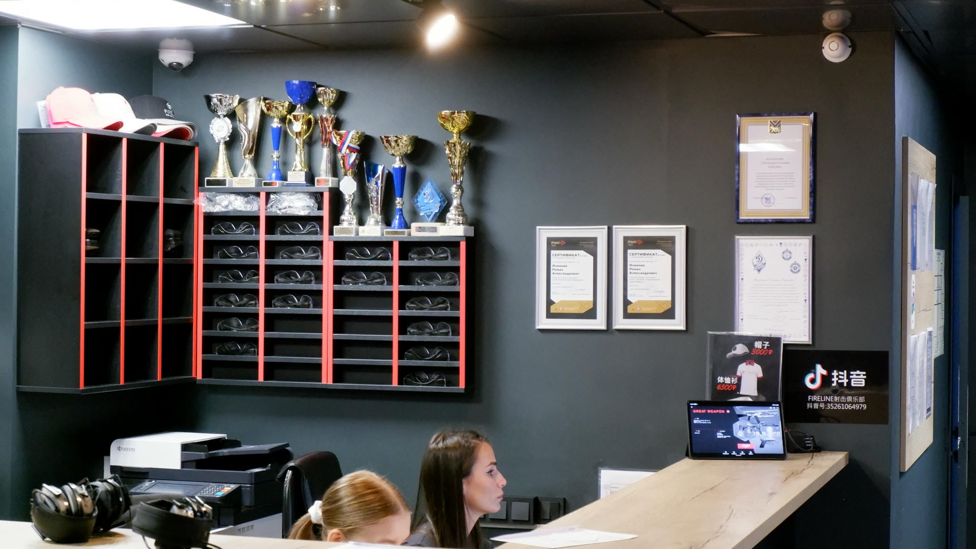 An indoor scene with many trophies and certificates hanging on the wall, two people behind a table with some equipment on it.
