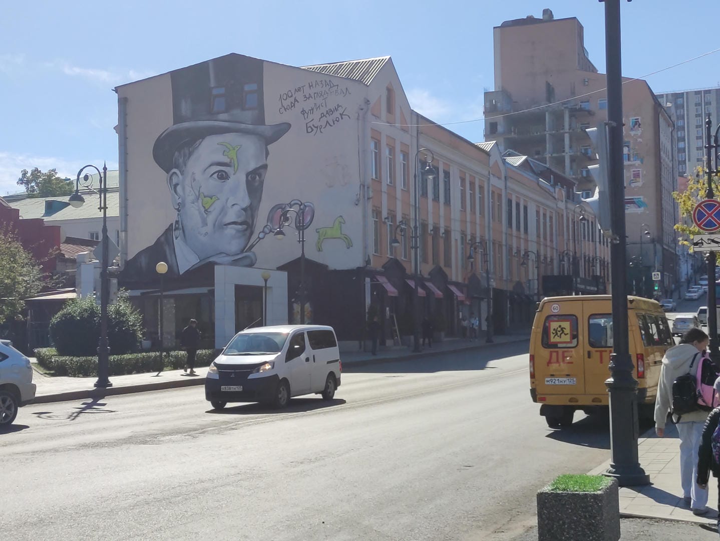 A large mural on the side of a building depicts a hat-wearing figure with Russian text beside it.