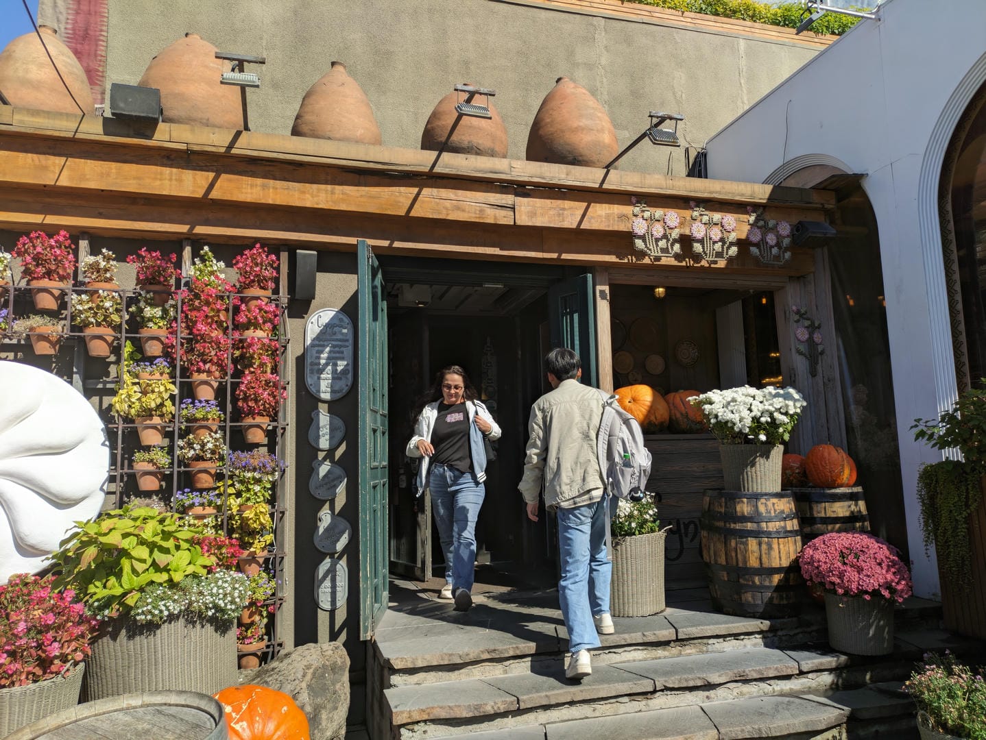 Two people are walking out of a doorway decorated with flowerpots and pumpkins, with clay pots hanging above.