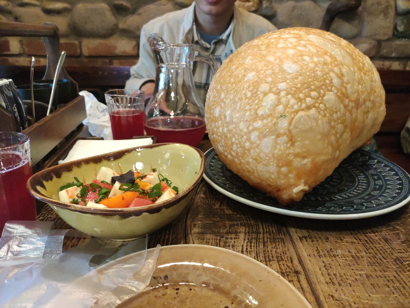 A large loaf of bread, a bowl of salad, and a glass of red beverage are on the dining table, with someone sitting in the background.