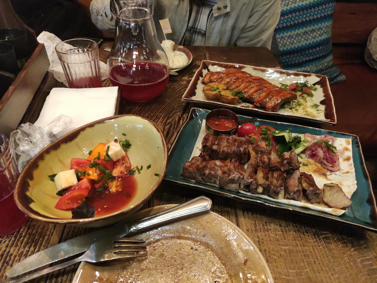 Grilled skewers, ribs, salad, and a glass of red beverage are neatly arranged on the dining table with utensils.