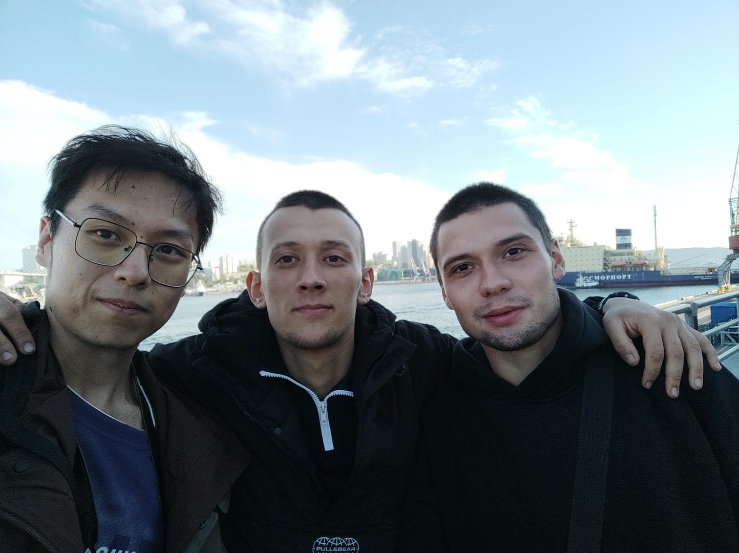 Three people stand outdoors for a group photo, with water and distant city buildings in the background.