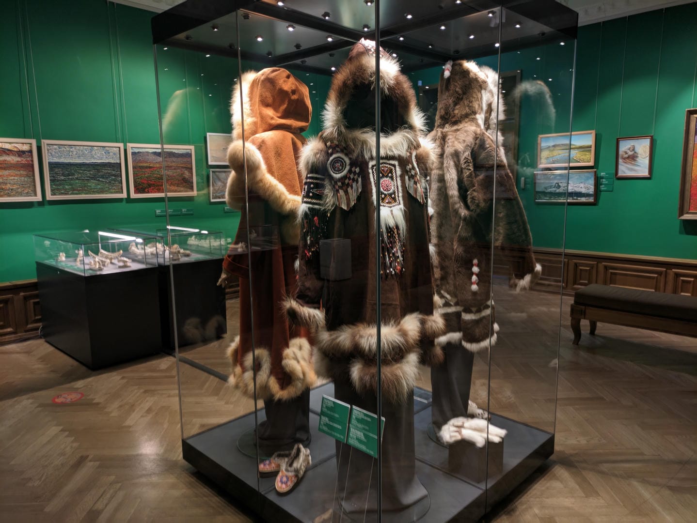 A museum display case contains three sets of traditional costumes, including fur coats and accessories.
