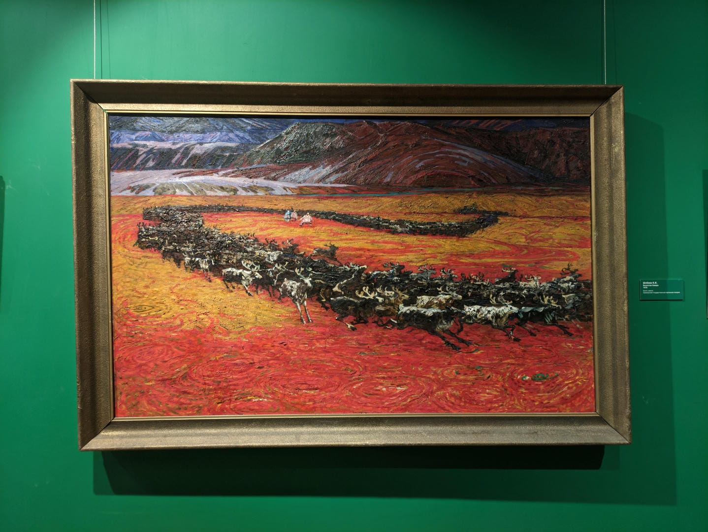 An oil painting in a frame depicts a herd of reindeer running across a red field with mountains in the background.