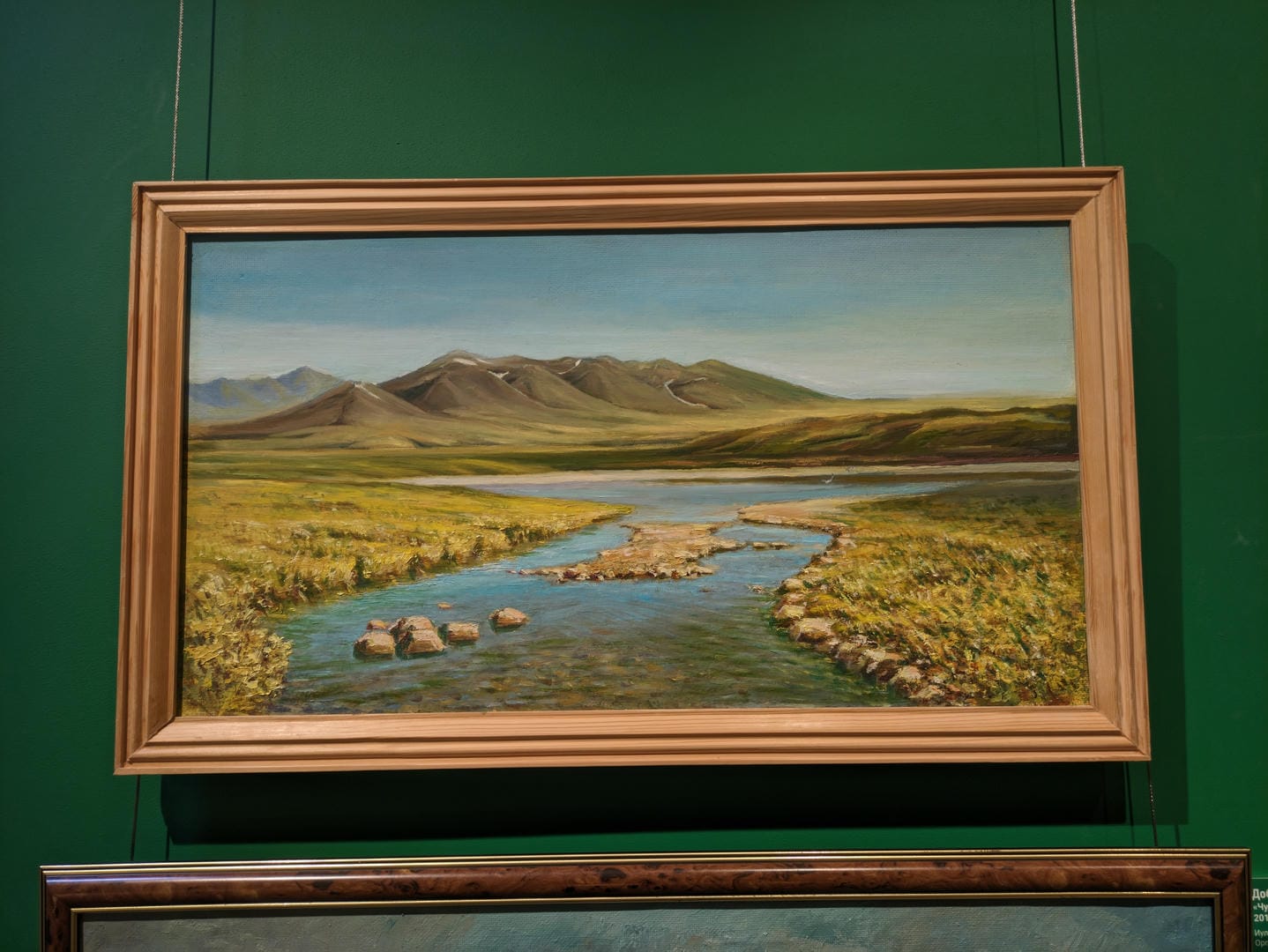 A painting shows a serene natural landscape, with a winding stream in the foreground and lush grasslands leading to rolling mountains.