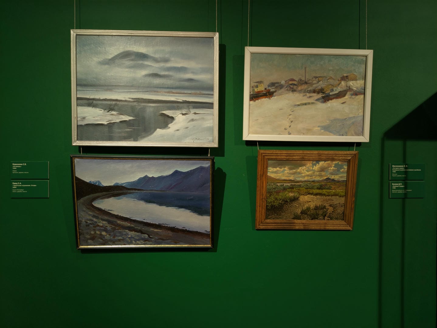 Four paintings hang on a green wall: the top-left depicts a river in the snow, the top-right shows a seaside village, the bottom-left presents a mountain river, and the bottom-right illustrates an oasis in the desert.