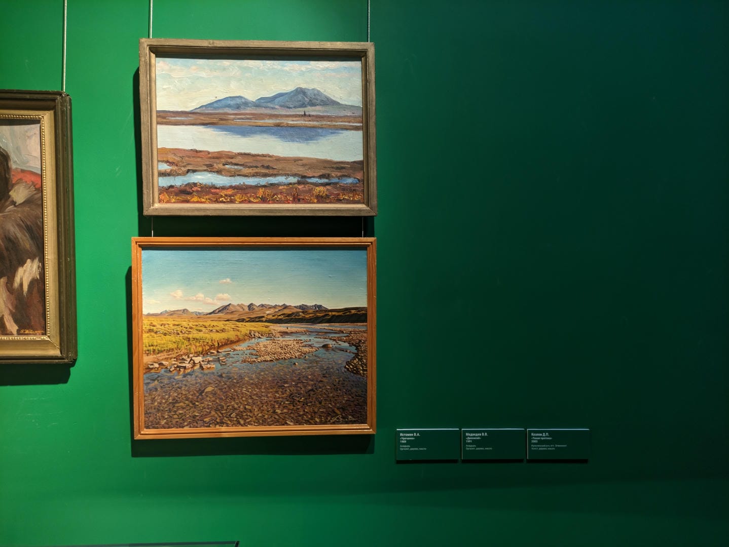 Three paintings hang on a green wall: the top-left depicts a lake with mountains, the top-right shows vast plains, and the bottom-left presents a mountain river.