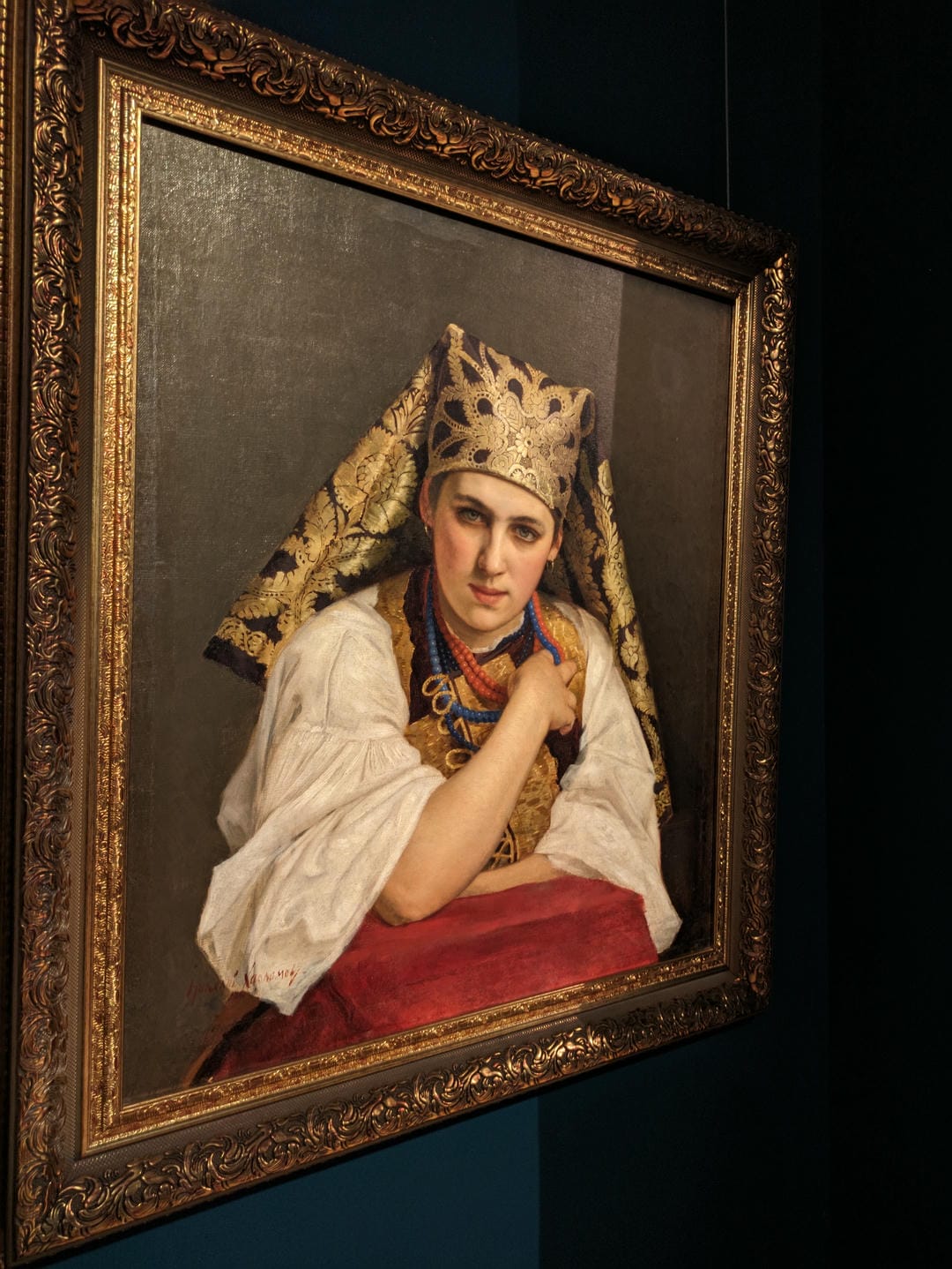 A woman in traditional attire sits before a red tablecloth, wearing an elaborately decorated headscarf.