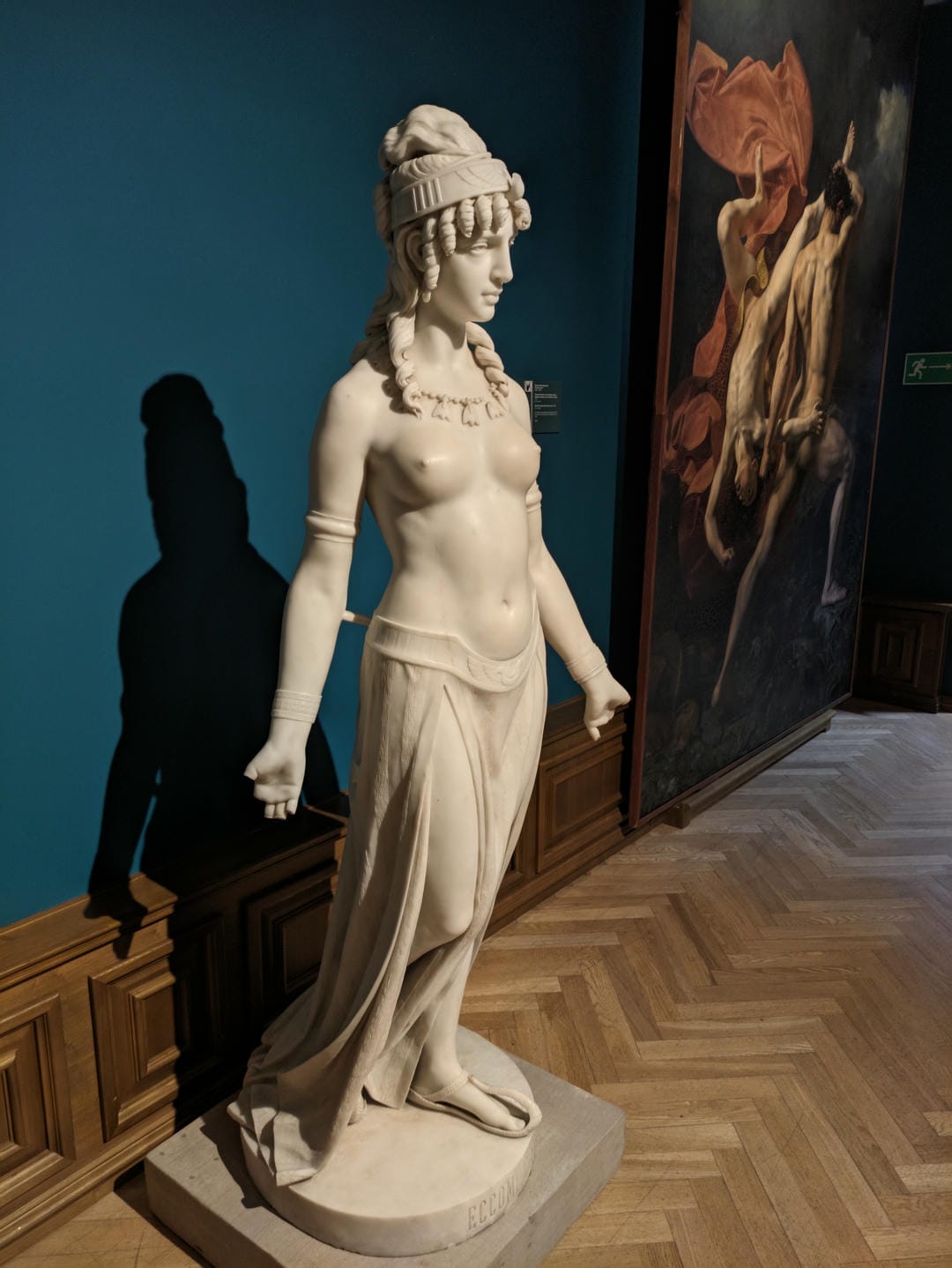A female statue stands bare-chested, wearing a long skirt, necklace, and headdress.
