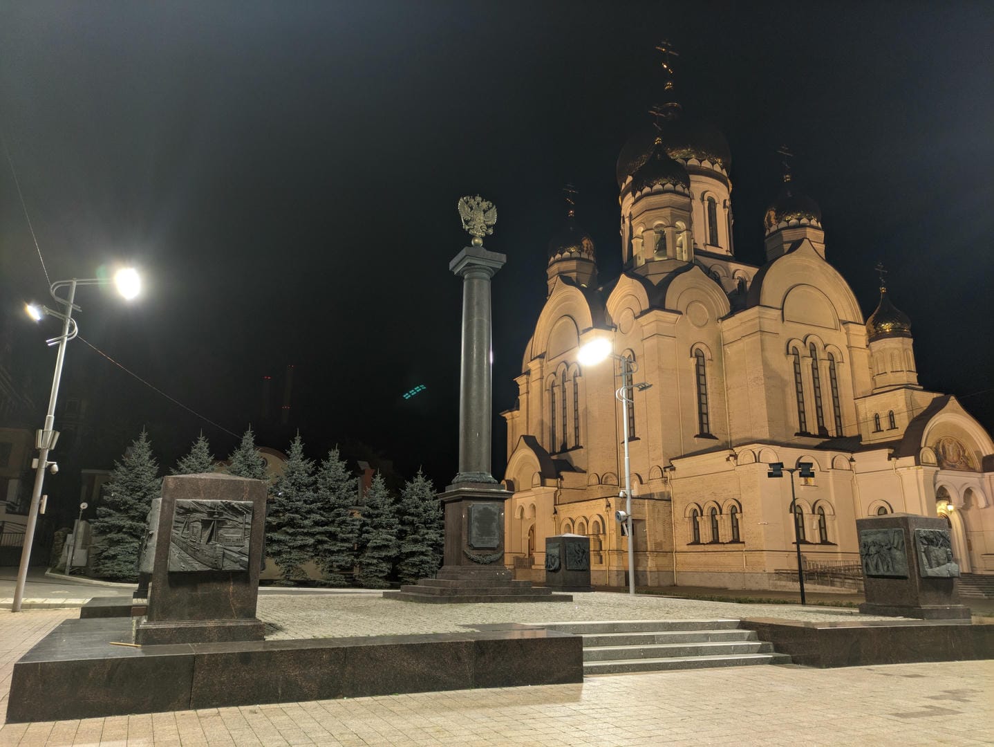 A nighttime square features a church, monument, and streetlights.