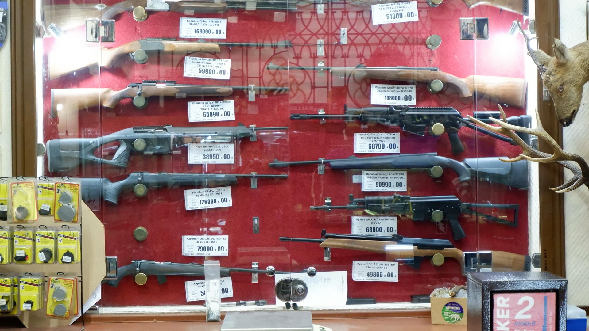 A display cabinet showcasing various firearms, each with a price tag below. A deer head specimen hangs on the right side.