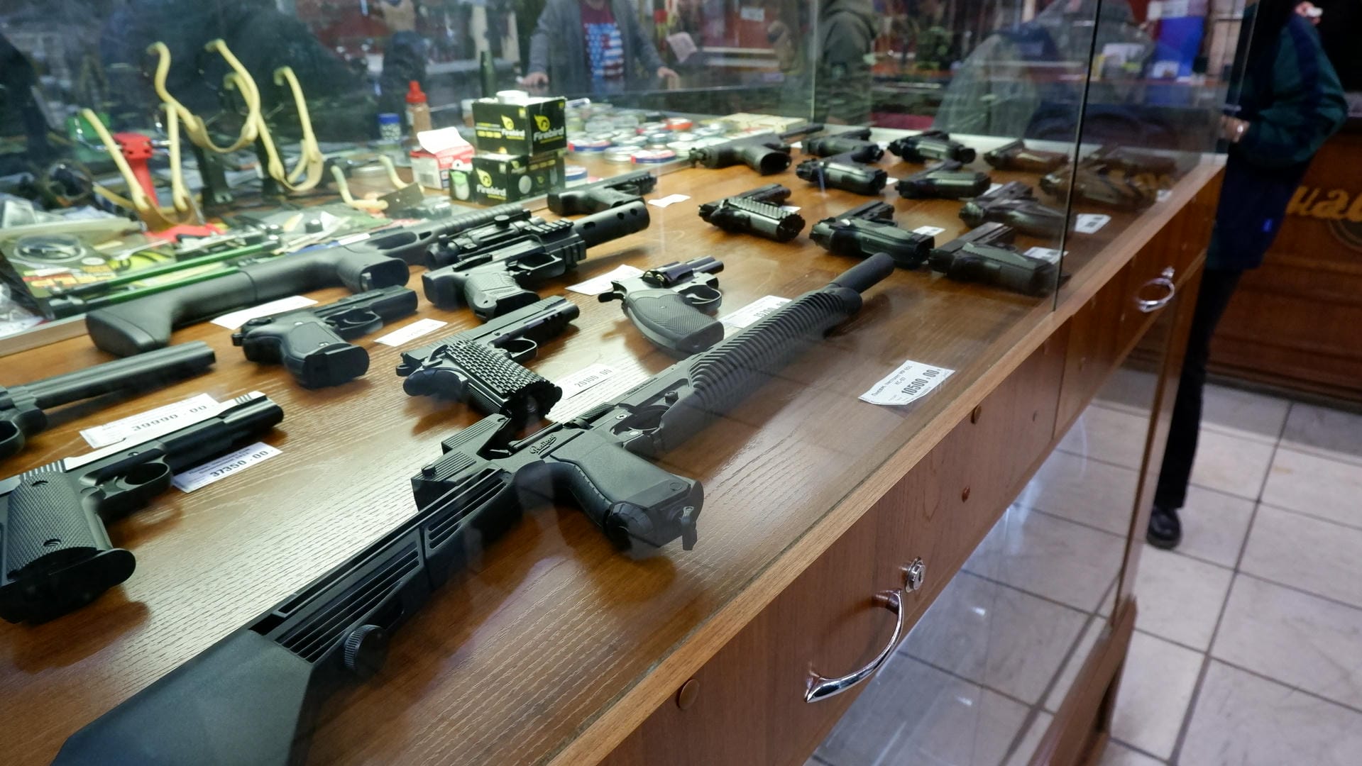 A display cabinet containing various firearms, each with a price tag beside it. There are small objects on the counter.