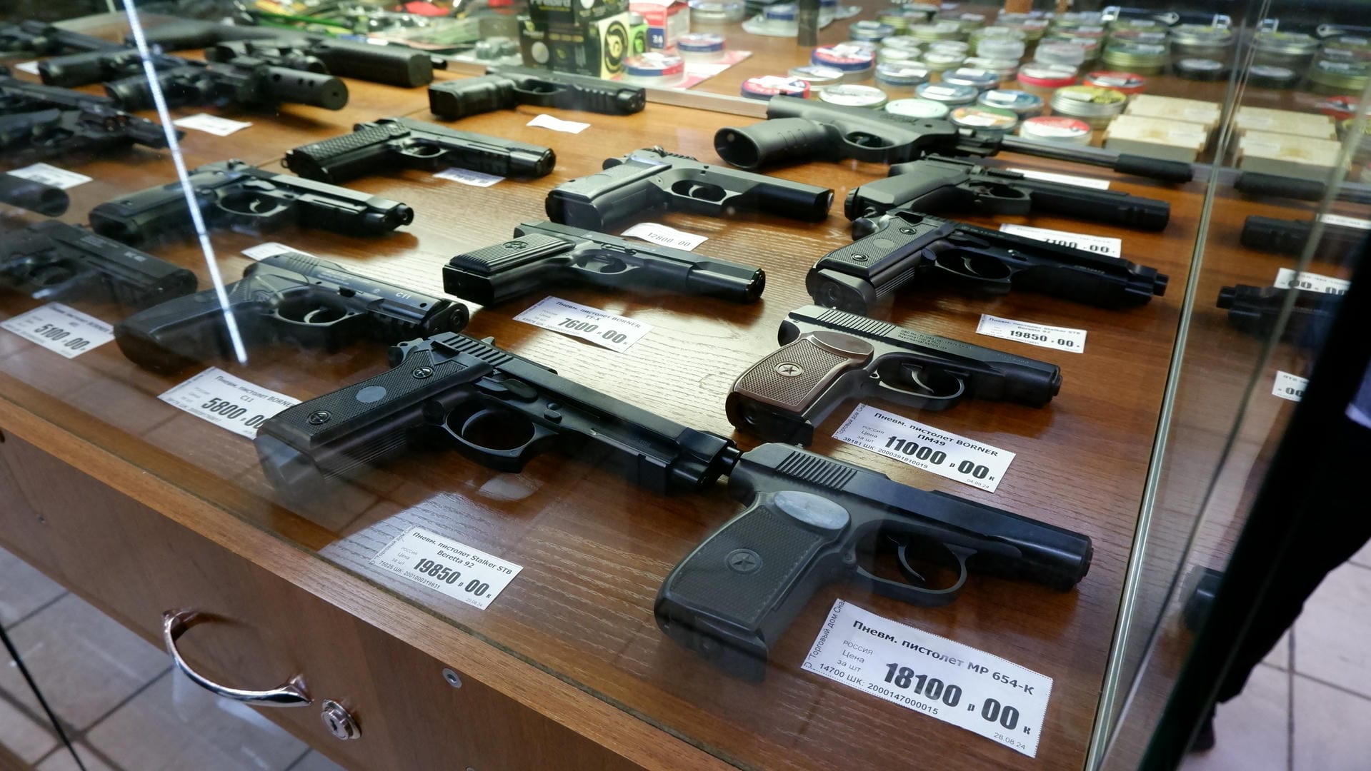A display cabinet showcasing various handguns, each with a price tag beside it. There are small objects on the counter.