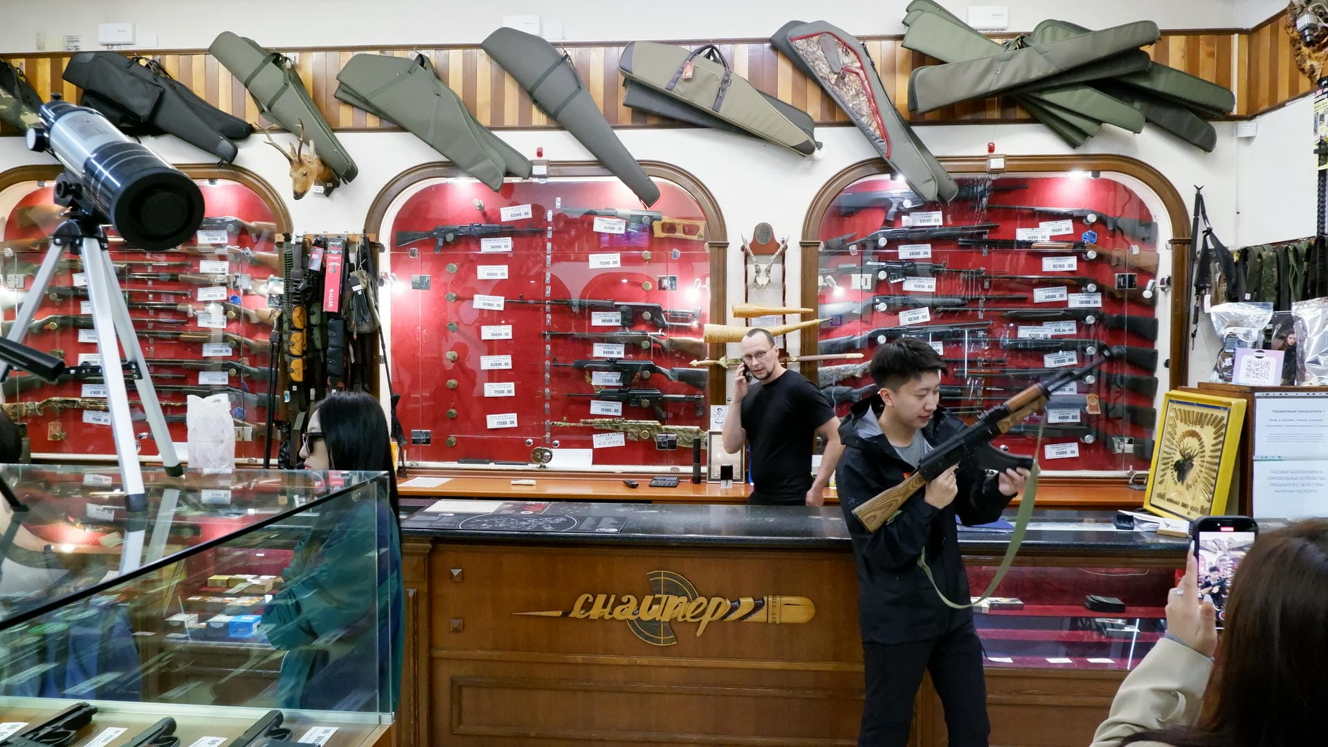 Many firearms hang on the walls inside the store. Two people stand in front of the counter, one holding a rifle. Gun bags hang from the ceiling.