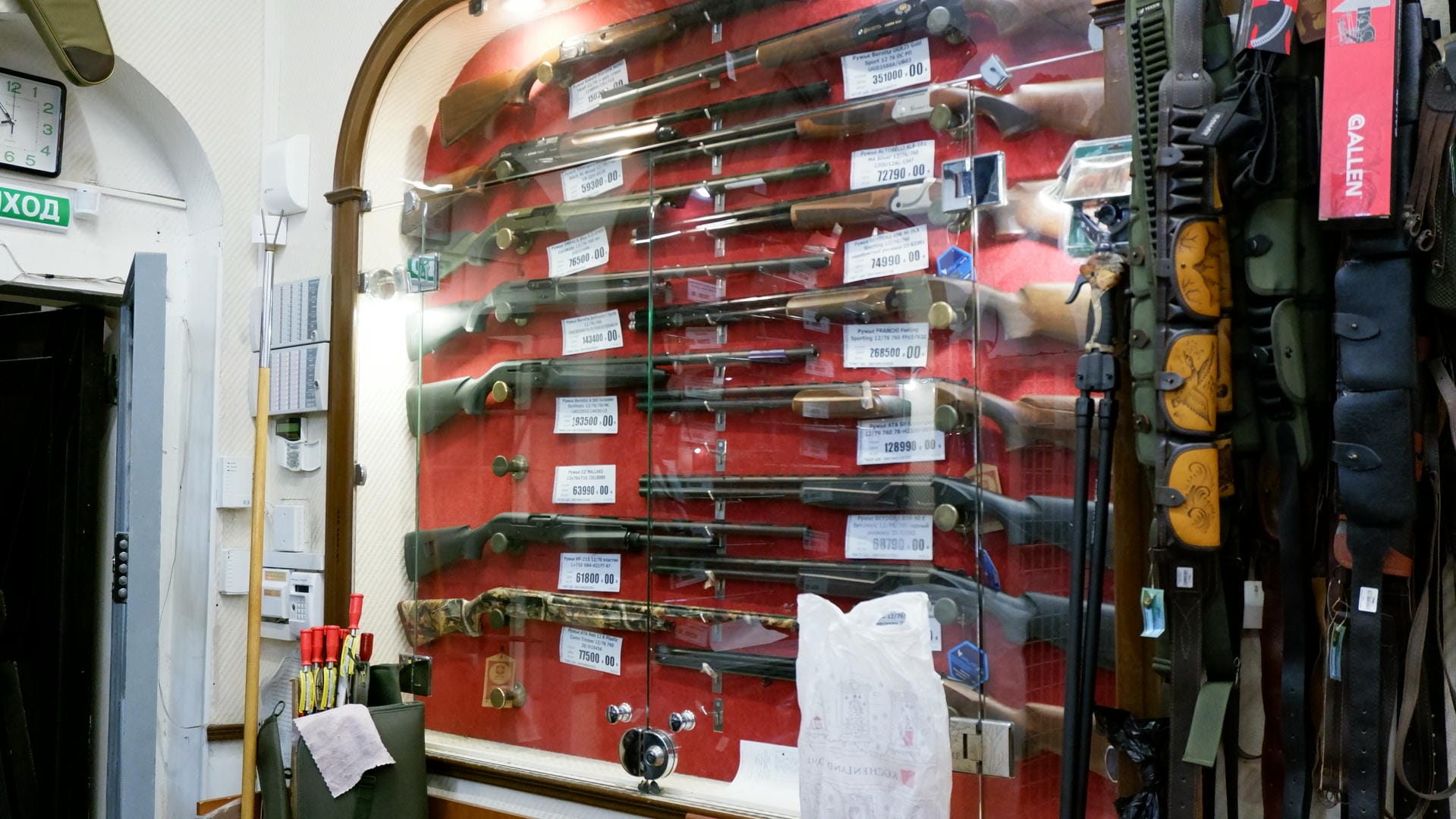 A display cabinet showcasing various firearms, each with a price tag below. Gun bags hang on the right side.