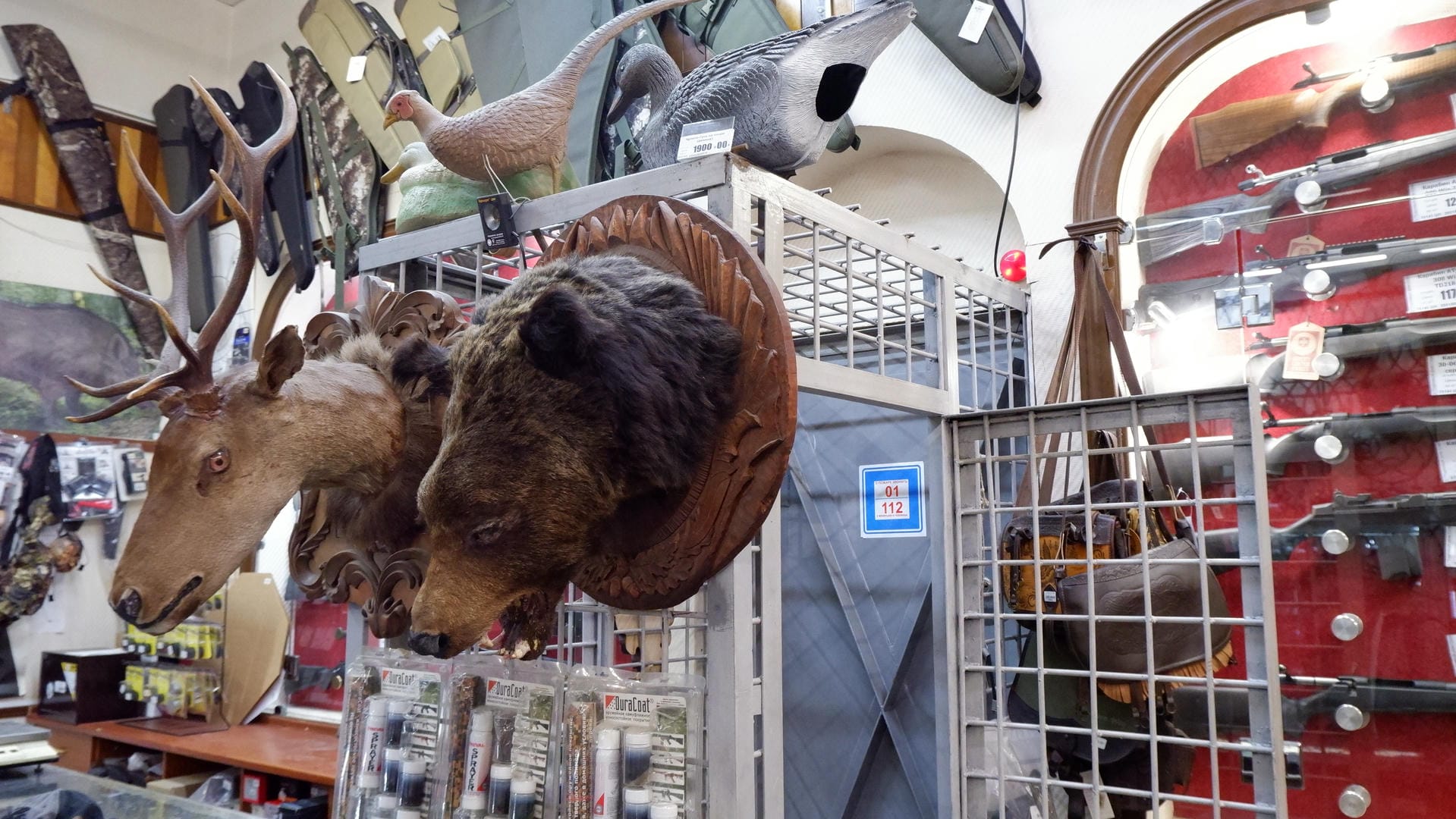 The store’s display cabinet contains various firearms, while animal specimens, including deer and bear heads, hang on the walls.