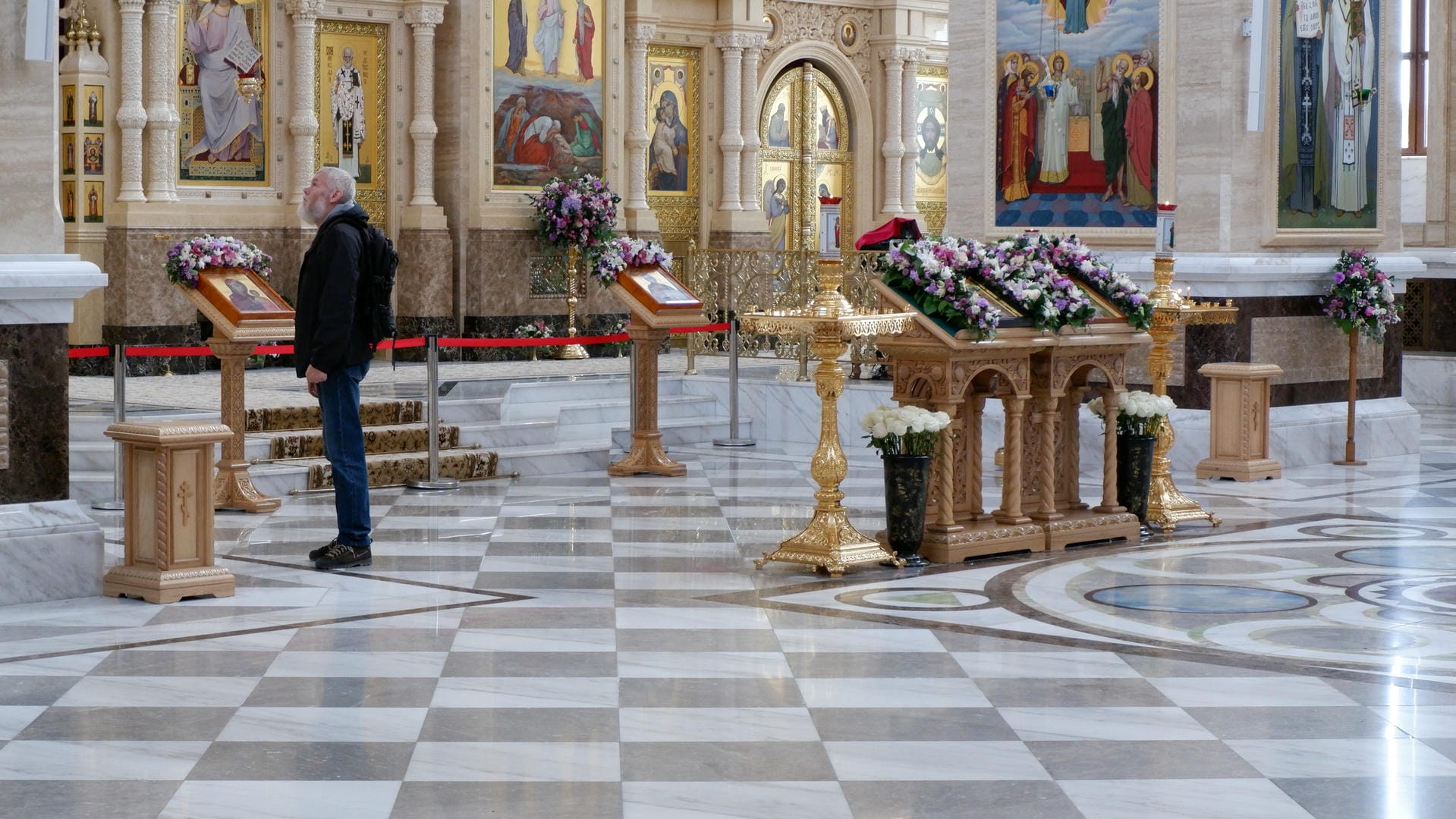 An elderly man stands inside the church, surrounded by flowers and religious paintings.