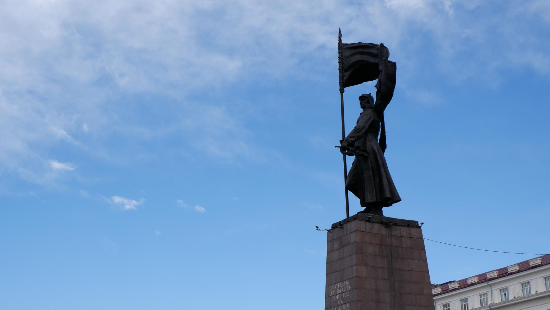 A statue stands on a pedestal, holding a flag, with a blue sky in the background.