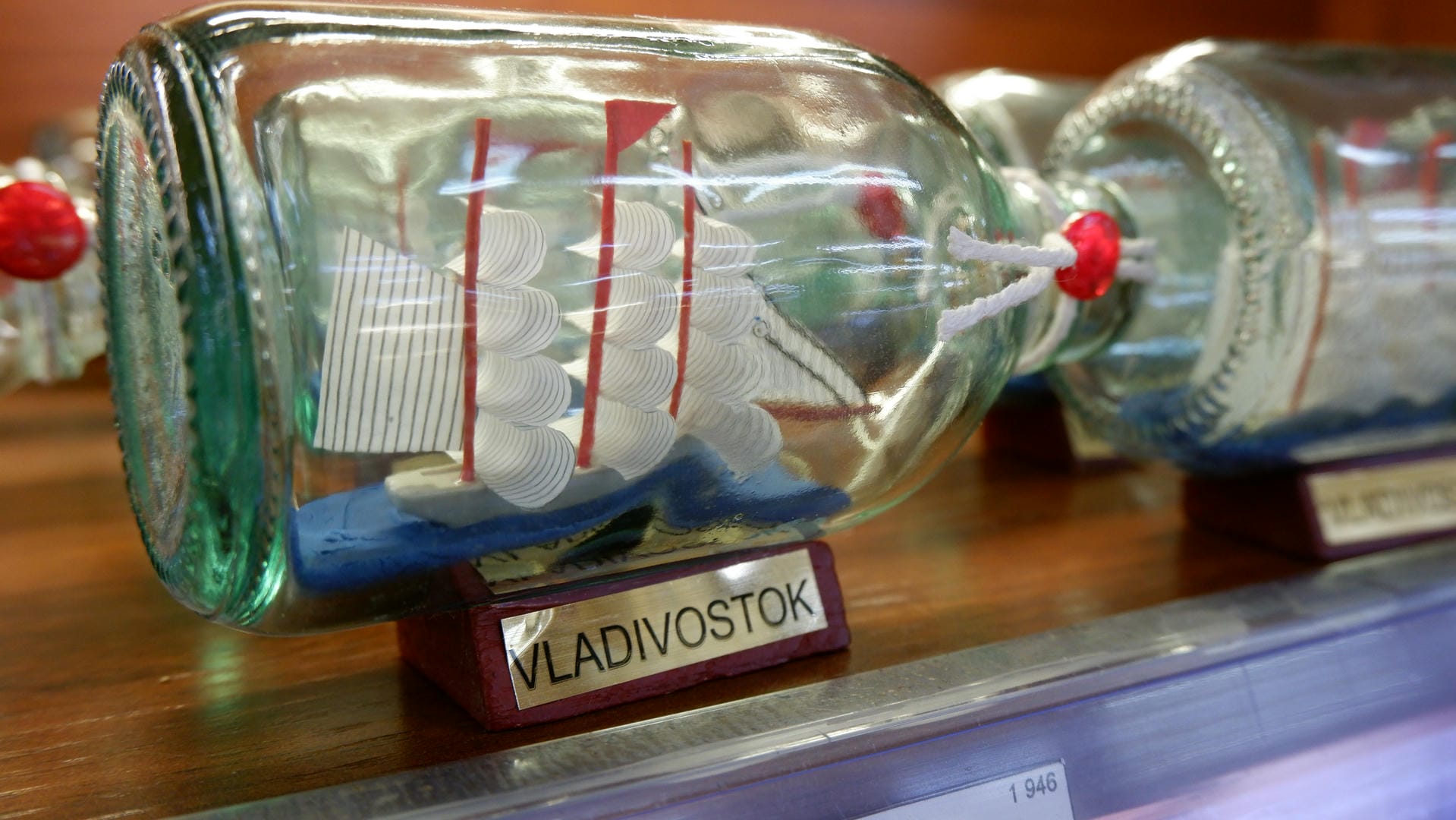 A glass bottle contains a model sailboat with red masts, sealed with a red stopper.
