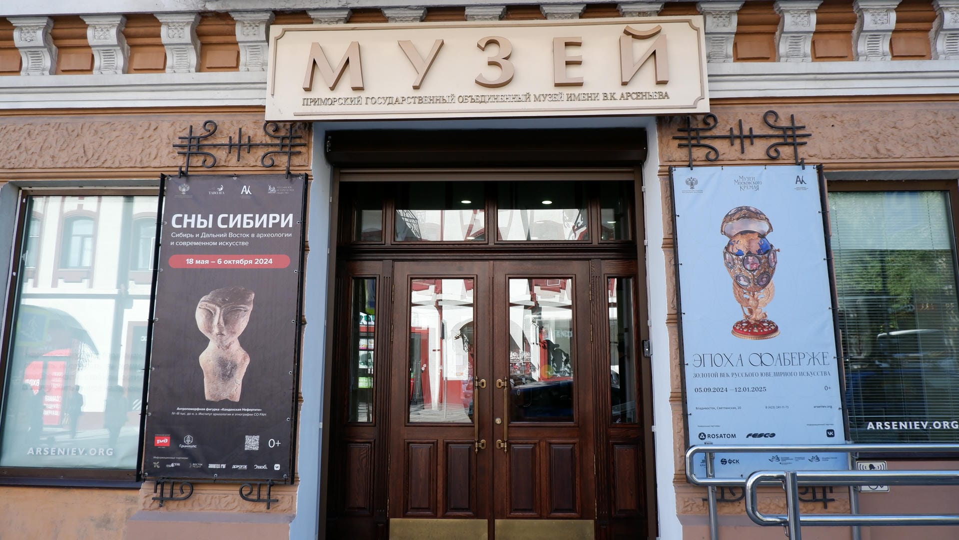 Above the museum entrance, the word “МУЗЕЙ” is displayed, with exhibition posters hanging on both sides.