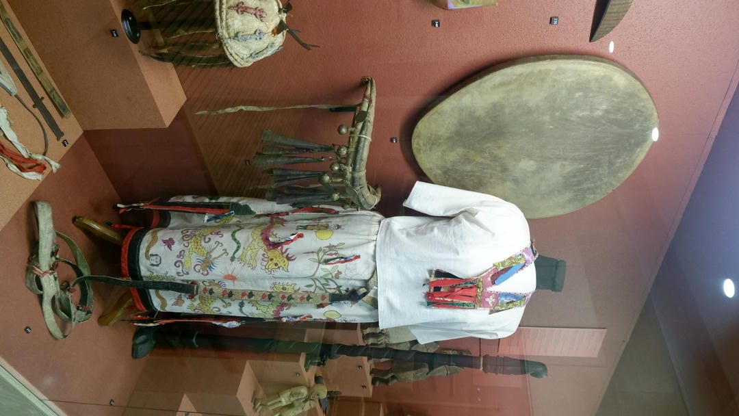 A set of traditional clothing is displayed on a wooden stand, accompanied by a round drum and other items.